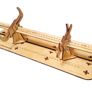 Puede incluir: Herramienta de madera con dos abrazaderas ajustables y una regla para medir y dar forma a objetos. La herramienta está hecha de madera marrón claro y tiene una regla marcada en centímetros.