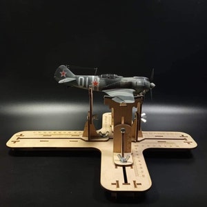 Peut inclure: Un support en bois pour modèle d'avion avec un modèle d'avion gris et blanc sur le dessus. L'avion a une étoile rouge sur la queue et le numéro "01" sur le côté. Le support a une règle de chaque côté et un bouton sur le devant.