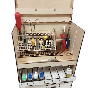 Może przedstawiać: Drewniany organizer na narzędzia z wieloma przegródkami i szufladami. Zawiera różne narzędzia, w tym śrubokręt z czerwoną rączką, nożyczki, szczypce i małe butelki. Organizer wykonany jest z jasnego drewna i ma pokrywę na zawiasach.