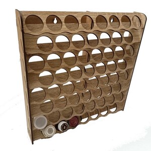 Könnte beinhalten: Ein hellbraunes Wandregal aus Holz mit einem Gitter aus kreisförmigen Aussparungen zur Aufbewahrung kleiner zylindrischer Behälter. Das Regal hat ein geneigtes Profil mit mehreren Reihen und Spalten, wobei vier kleine Behälter eingesetzt sind.