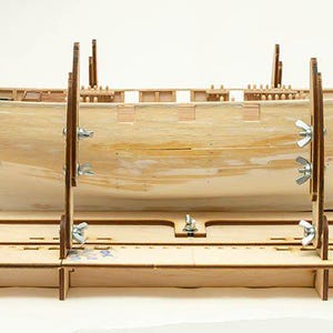 Op de afbeelding: Een houten modelboot in aanbouw, vastgehouden door een houten frame met verstelbare steunen. De onafgewerkte romp is lichtbruin, met zichtbare interne structuren. Het frame heeft meerdere niveaus en metalen bevestigingen.
