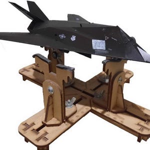 May include: A black model airplane, resembling a stealth fighter jet, is displayed on a wooden stand. The model has detailed wings and tail fins, with small decals. The stand is made of light brown wood, with adjustable supports and wing nuts.