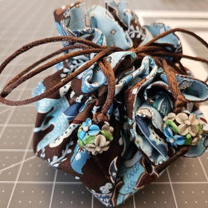 May include: A blue and brown drawstring pouch with a paisley pattern. The pouch is closed with a brown cord and has blue and white beads on the ends of the cord.
