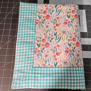 May include: A turquoise and white checkered fabric with a pink and yellow floral print in the center. The fabric is folded in half and the edges are sewn.