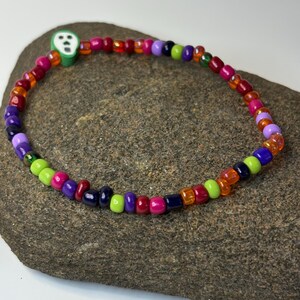 May include: A colorful beaded bracelet with a variety of small, round beads in shades of red, orange, purple, green, and pink. A green alien-shaped charm is attached to the bracelet. The bracelet is displayed on a gray stone surface.