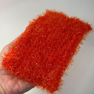 Puede incluir: Artículo rectangular de ganchillo de color naranja brillante con un aspecto texturizado y difuso. El artículo se sostiene en una mano y parece ser un paño de lavado o una almohadilla de limpieza hechos a mano. El color es un naranja vibrante y sólido con un ligero brillo metálico.