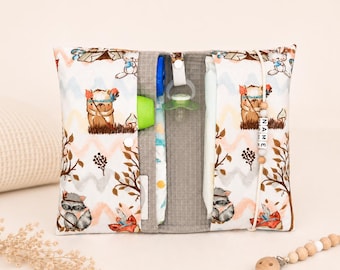 May include: A white and gray diaper clutch with a woodland animal print. The clutch is open, revealing a pacifier, diaper, and other baby essentials. A wooden pacifier clip with the name "NAME" is attached.