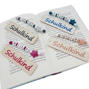 May include: Wooden bookmarks with the word "Schulkind" in various colors, accented with beaded strings and star-shaped charms. The bookmarks are displayed open in a book, with the text "Schulkind" in different colors.