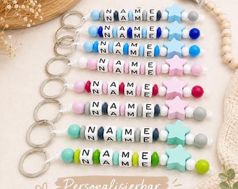 May include: A collection of personalized baby pacifier clips with the name "NAME" spelled out in white letter beads. Each clip features a different color scheme, including blue, pink, and green, with star-shaped wooden accents. The text "Personalisierbar" is visible.