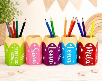 May include: Five hexagonal pencil holders in green, pink, purple, blue, and red, each topped with a light yellow rim. The holders feature white star icons and personalized names or text including Schulkind 2026, Sina, Lena, Lenni, and Mila.