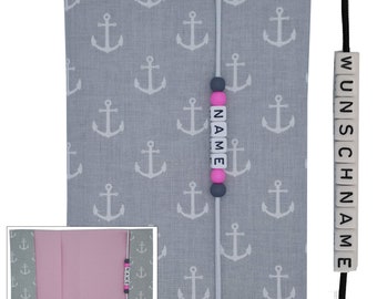 May include: A gray fabric book cover with a white anchor pattern. A white string with black and pink beads spells out the word "NAME". A separate black string with white beads spells out the word "WUNSCHNAME".
