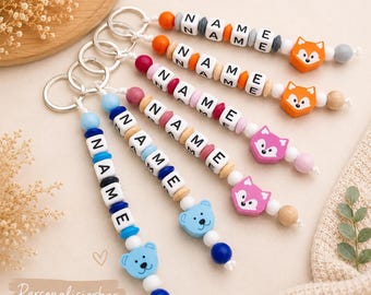 May include: A collection of personalized keychains with the word "NAME" spelled out in wooden letter beads. The keychains feature colorful beads, fox and bear charms, and silver key rings. The text "Personalisierbar mit deinem Wunschnamen" is visible.
