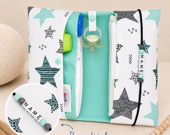 May include: A white and teal star-patterned baby changing clutch with a teal interior. The clutch contains a diaper, pacifier, and other baby essentials. A personalized pacifier clip with the name "NAME" is attached. The text "Personalisierbar" is visible.
