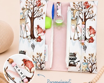 May include: A baby changing pad with a woodland animal print. The pad is open, revealing pockets for diapers, a pacifier, and a bottle. A wooden pacifier clip with the name "NAME" is attached. The text "Personalisiert" is visible.