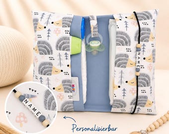 May include: A baby travel organizer with a white background and a pattern of hedgehogs, trees, and mushrooms. The organizer has a blue interior and holds a pacifier, bottle, and other baby essentials. The text "Personaliserbar" is visible.