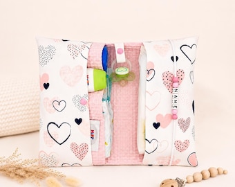 May include: A white and pink baby diaper bag with a heart pattern. The bag is open, revealing diapers, a pacifier, and a tube of cream. A wooden pacifier clip with wooden beads is also visible.