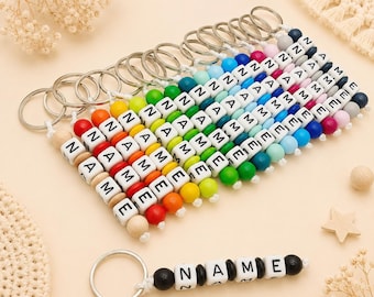 May include: A collection of colorful beaded keychains spelling out names. Each keychain features wooden letter beads and vibrant accent beads in a rainbow of colors. One keychain spells "NAME" with black beads. The keychains are attached to silver key rings.