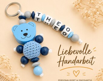 May include: A light blue wooden bear keychain with a crocheted body and dark blue wooden bead limbs. The keychain includes a beaded strand spelling "THEO". The image also includes the text "Liebevolle Handarbeit".