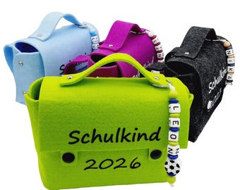 May include: A collection of felt satchels in light blue, purple, green, and charcoal gray. Each bag has a handle and a clasp closure. The green bag has the text "Schulkind 2026" and a beaded charm. The other bags have beaded charms.