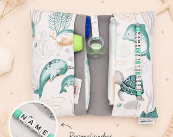 May include: A baby changing pad with a marine life pattern, featuring whales, crabs, and dolphins in teal and gray. Includes a pacifier, diaper, and a personalized name bead chain. The text "Personalizable" is also visible.