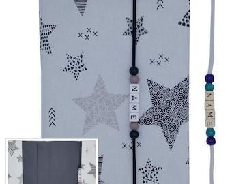 May include: A gray and white fabric book cover with a black star pattern. The cover has a black strap with wooden beads that spell out the word "NAME".