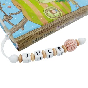 May include: A handmade bookmark with the name "JULE" spelled out in white letter beads. The bookmark features wooden beads, a crocheted peach-colored ball, and a white cord attached to a children's book.