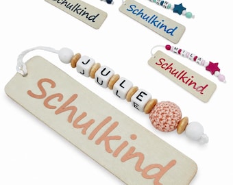 May include: Wooden bookmarks with the word "Schulkind" printed in various colors. Each bookmark has a beaded charm with a name spelled out in letter beads. The charms include star and round beads.