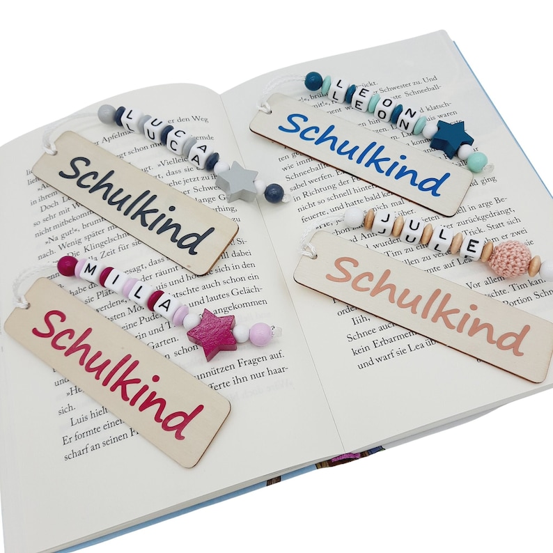 May include: Wooden bookmarks with the word "Schulkind" in various colors, accented with beaded strings and star-shaped charms. The bookmarks are displayed open in a book, with the text "Schulkind" in different colors.