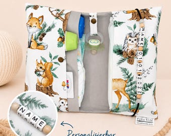 May include: A white and gray baby changing pad with a woodland animal print, including foxes, squirrels, and deer. The pad is open, revealing pockets for diapers, a pacifier, and other baby essentials. A wooden bead chain spells out "NAME".