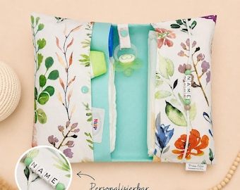 May include: A floral-patterned baby changing clutch with a teal interior, holding a diaper, pacifier, and other baby essentials. The clutch features a watercolor design of leaves and flowers in shades of green, purple, and orange. The text "NAME" is visible.