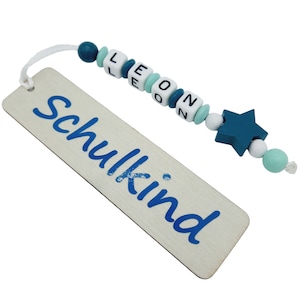 May include: A wooden bookmark with the word "Schulkind" in blue script. A string of beads spells out the name "LEON" in white and blue. A teal star and beads complete the design. The bookmark is a light wood color.