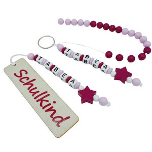 May include: A collection of handmade wooden accessories in shades of pink and white. Includes a bookmark with the word "Schulkind", beaded keychains spelling "TABEA", and star-shaped charms. The beads are strung on white string.