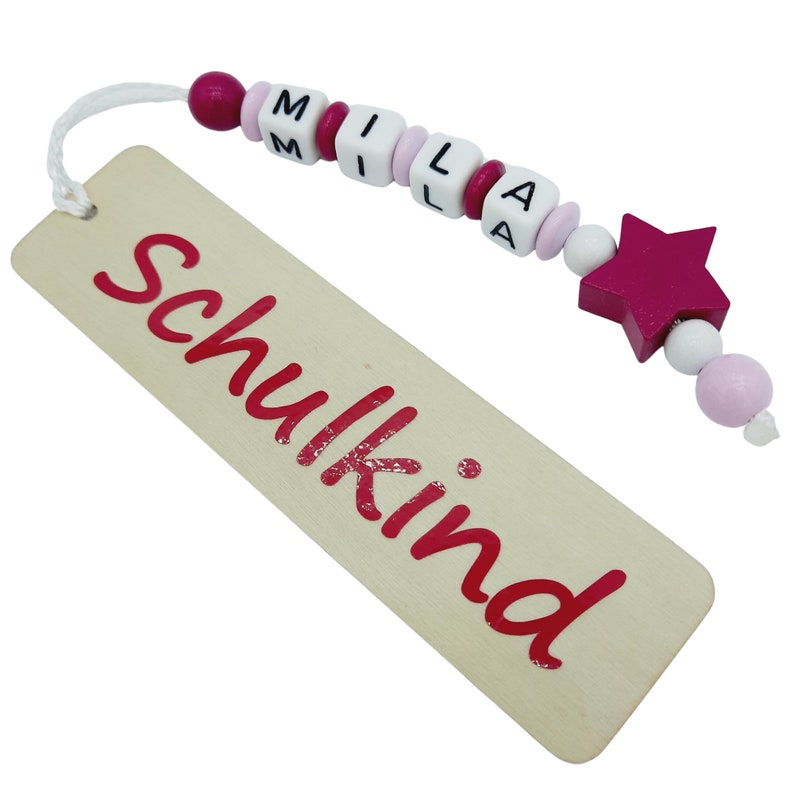 May include: A wooden bookmark with the word "Schulkind" in red script. The bookmark is attached to a string of beads spelling "MILA" and a magenta star-shaped bead.