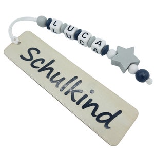 May include: A wooden bookmark with the word "Schulkind" printed in black. The bookmark is attached to a string of beads spelling "LUCA", with a gray star-shaped bead at the end. The beads are white, gray, and navy blue.
