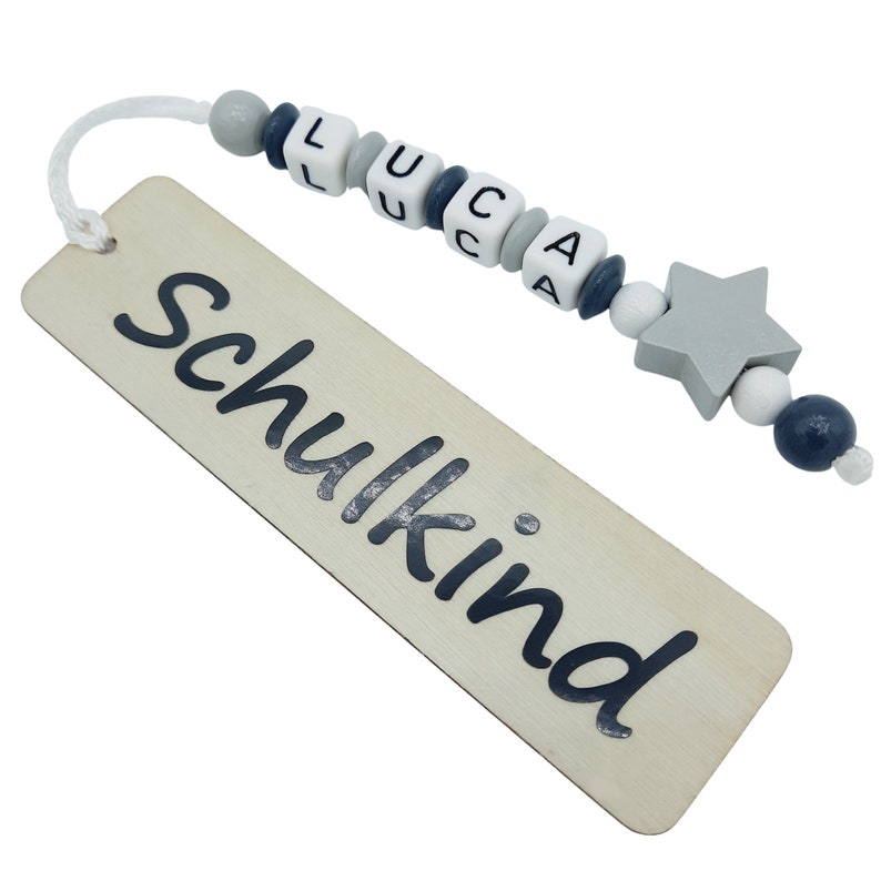 May include: A wooden bookmark with the word "Schulkind" printed in black. The bookmark is attached to a string of beads spelling "LUCA", with a gray star-shaped bead at the end. The beads are white, gray, and navy blue.