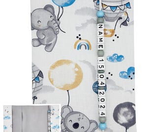May include: A white fabric baby book cover with a whimsical print of elephants, hot air balloons, and balloons in shades of gray, blue, and yellow. A beaded string with the name and date is attached. The cover has a gray interior.