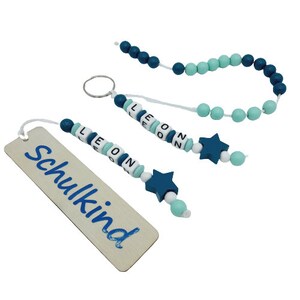 May include: A collection of wooden bead accessories in shades of blue and white. Includes a bookmark with the word "Schulkind", a keychain with the name "LEON", and a beaded strand. Each item features star-shaped accents.