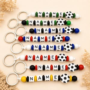 Könnte beinhalten: Eine Sammlung personalisierter Schlüsselanhänger mit dem Wort "NAME" in weißen Buchstabenperlen. Jeder Schlüsselanhänger hat einen Fußball-Charm und farbige Perlen in verschiedenen Kombinationen aus Grün, Schwarz, Blau, Rot und Gelb.