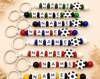 May include: A collection of personalized keychains with the word "NAME" spelled out in white letter beads. Each keychain features a soccer ball charm and colorful beads in various combinations of green, black, blue, red, and yellow.