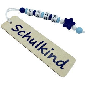 May include: Wooden bookmark with the word "Schulkind" in dark blue script. The bookmark is attached to a string of beads spelling "LARS" and a blue star-shaped bead. The beads are white, light blue, and dark blue.