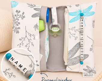 May include: A fabric diaper clutch featuring a white background with gray botanical illustrations and bright blue dragonfly motifs. The interior contains a pacifier and wipes. A white cord with gray and blue beads displays the text NAME 01 12 2022.