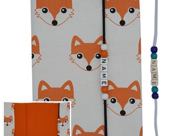 May include: A white fabric book cover with a fox pattern and a black bookmark with white beads that spell "NAME". The cover has a pocket on the front with a matching fox pattern. A white string with blue and turquoise beads that spell "NAME" is attached to the cover.