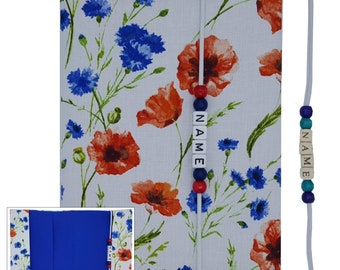 May include: A floral journal cover with a white background, featuring red poppies and blue cornflowers. The journal has a blue interior and a matching elastic closure with the word "NAME" spelled out in wooden beads.