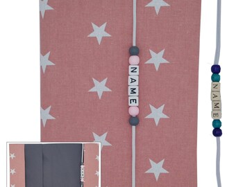 May include: A pink fabric book cover with white stars and a white cord with a name bead set that spells "NAME" in black letters. The beads are pink, gray, and white. The cord is attached to a second set of beads that spell "NAME" in black letters. The beads are blue, teal, and gray.