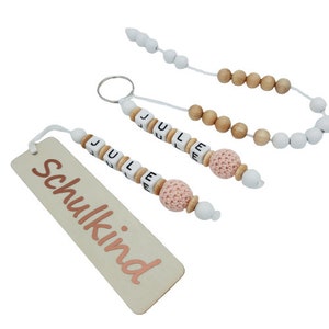 May include: A collection of personalized accessories, including a wooden bookmark with the word "Schulkind" in rose gold, a keychain, and a beaded strand. The keychain and strand feature the name "JULE" spelled out with lettered beads, and a pink crocheted bead.