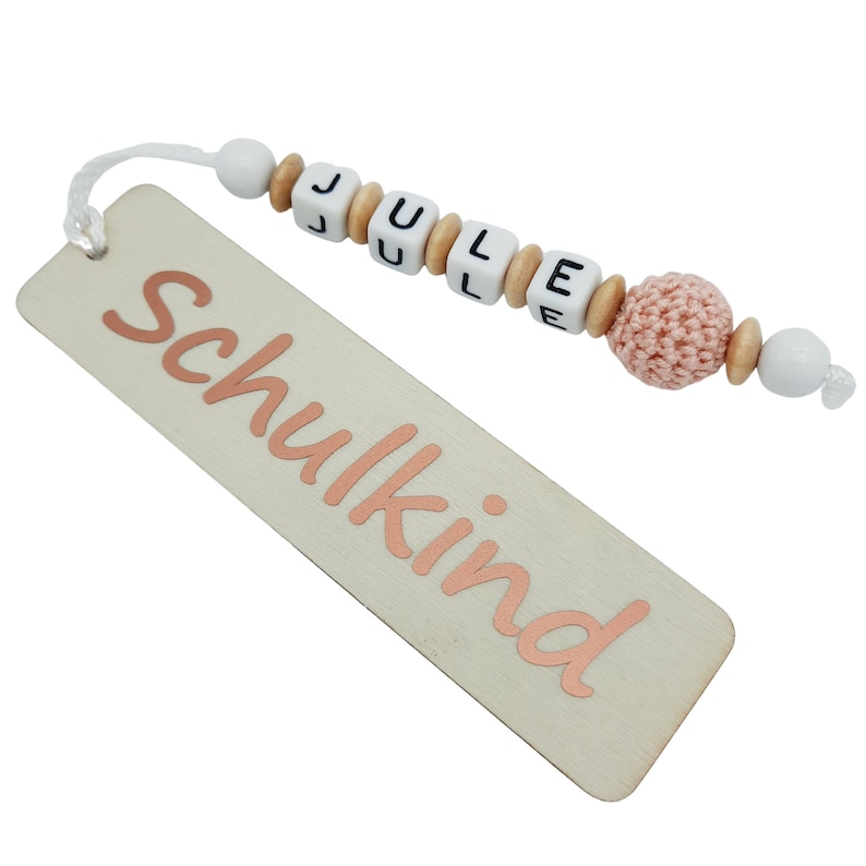 May include: A wooden bookmark with the word "Schulkind" in rose gold script. The bookmark is attached to a string of white beads, wooden beads, and lettered beads spelling "JULE", and a pink crocheted bead.