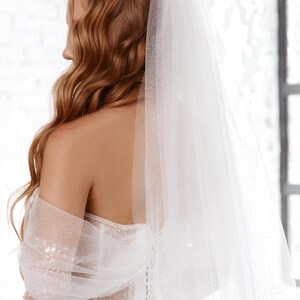 May include: A bride in a white wedding dress with a sheer, sparkling veil. The dress has a detailed bodice and a flowing skirt. The bride's auburn hair is styled in loose waves, and a sheer, embellished wrap covers her shoulders.