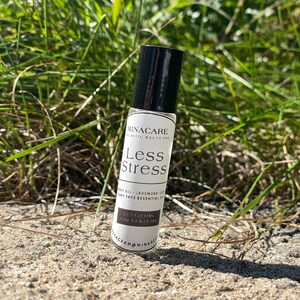 May include: A small, white bottle with a black cap and a rollerball applicator. The bottle is labeled "MINACARE Holistic Selfcare Less Stress" and "Connecting You To Nature".