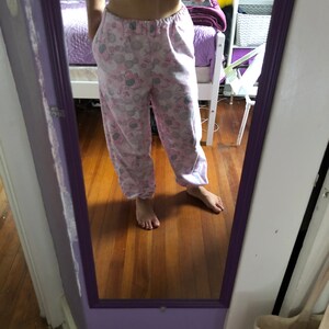 May include: Pink and grey pyjama trousers with a cartoon print of a character wearing a crown. The trousers have a drawstring waistband and pockets.