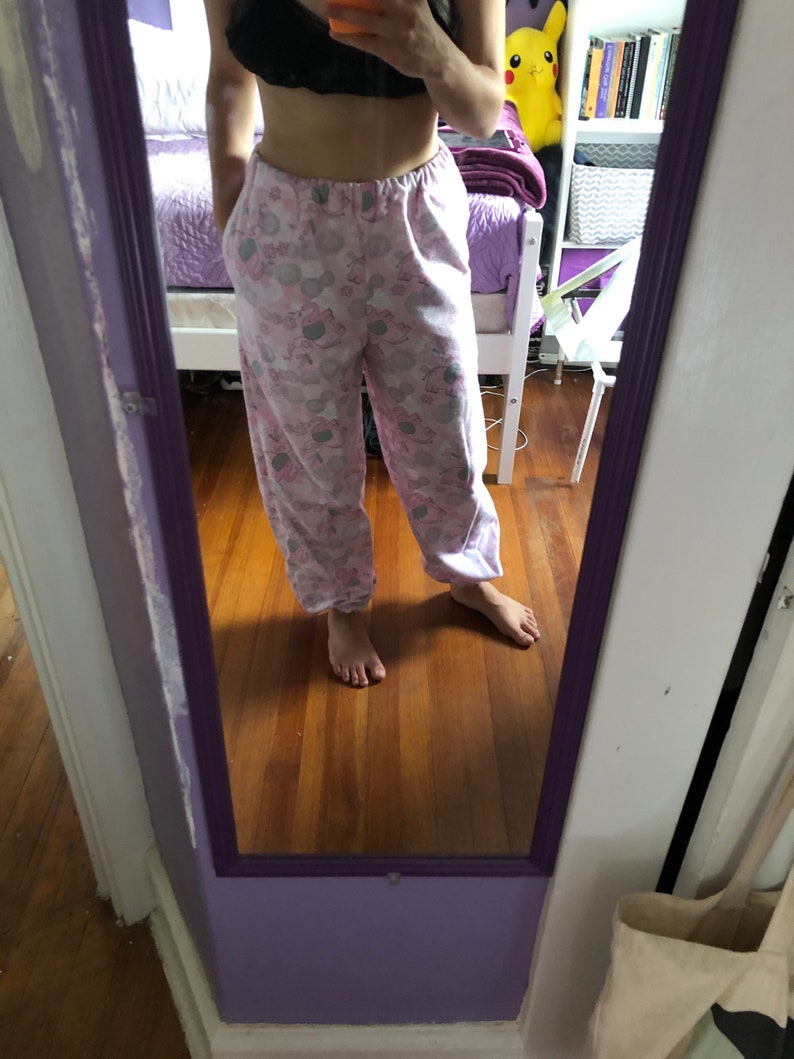 May include: Pink and grey pyjama trousers with a cartoon print of a character wearing a crown. The trousers have a drawstring waistband and pockets.